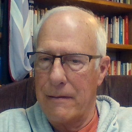 Charles Schuster sitting in a chair with a blue sweatshirt and books in the background