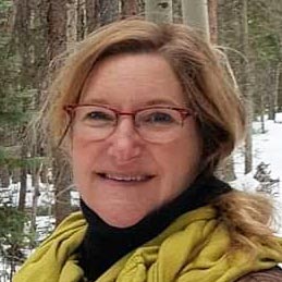 Close-up photo showing smiling face and the shoulder area of Rebecca McFee, with snowy hillside and tall lodgepole pines in the background.